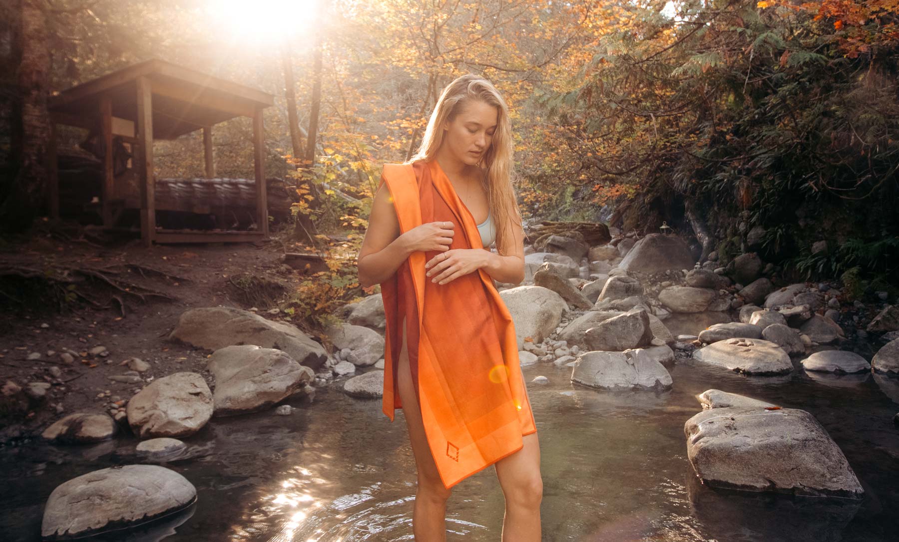 Woman holding an orange towel in a shallow forest stream near a cabin