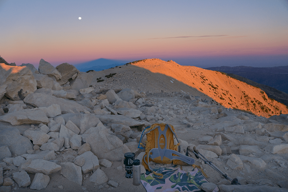 Hiking gear rests on rocky mountain terrain at sunset with distant peaks and a visible moon