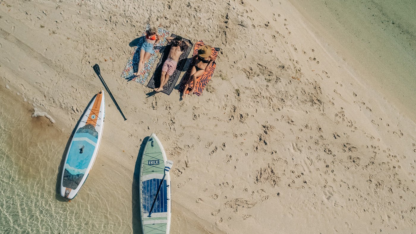 People relaxing on patterned beach towels beside paddleboards on a sandy beach
