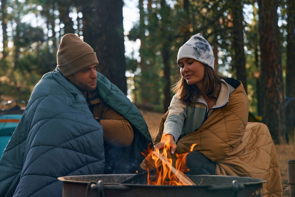 Two people wrapped in puffer blankets sit beside a campfire in a forest