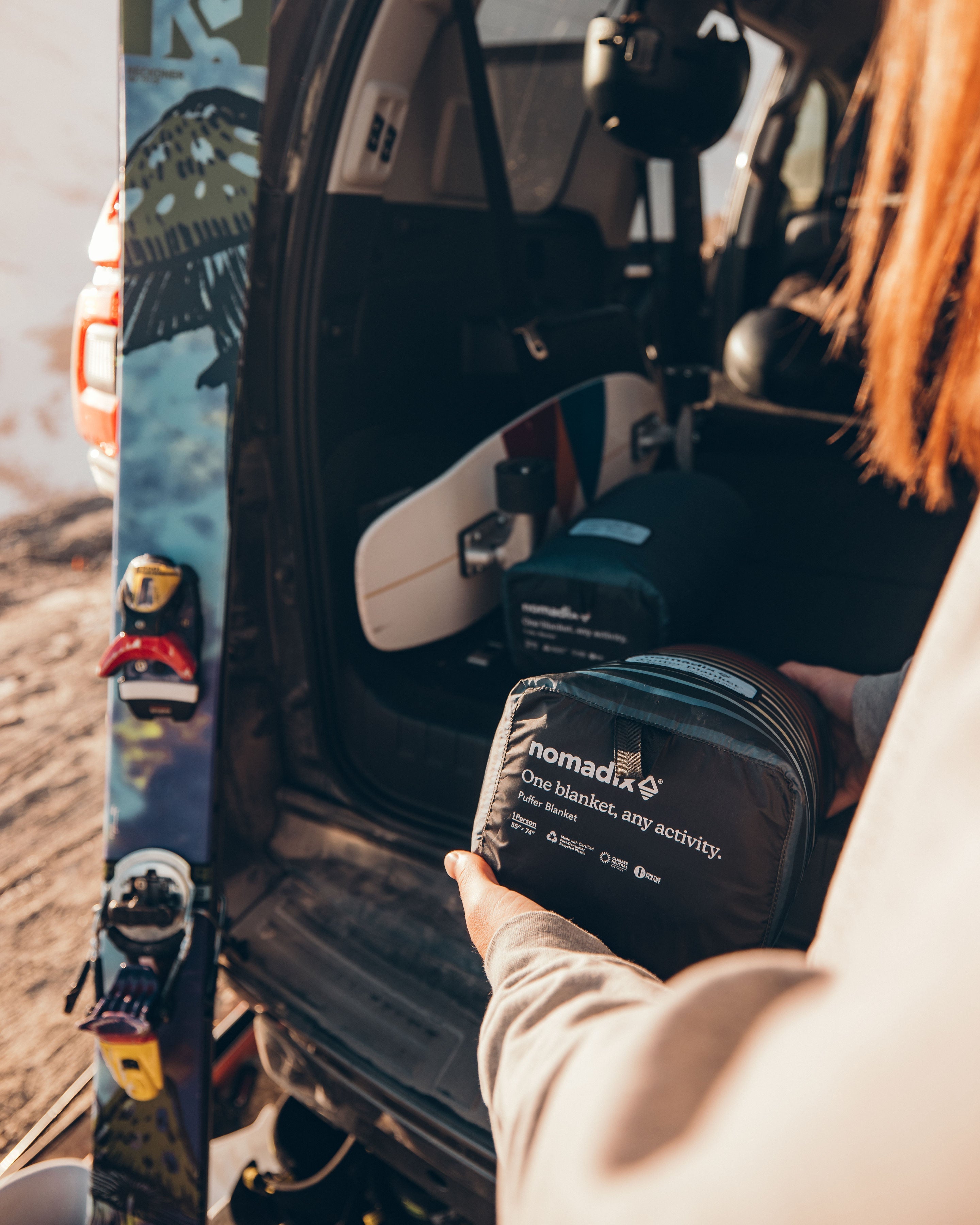 Person placing a Nomadix Puffer Blanket in an SUV beside snowboards