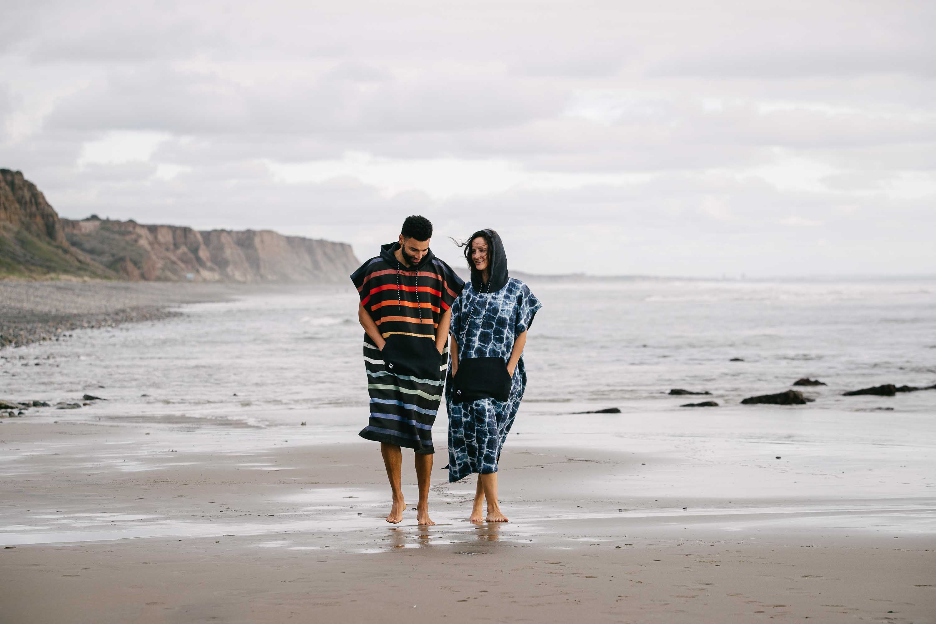 Two people wearing changing ponchos walk barefoot along a beach beside the ocean