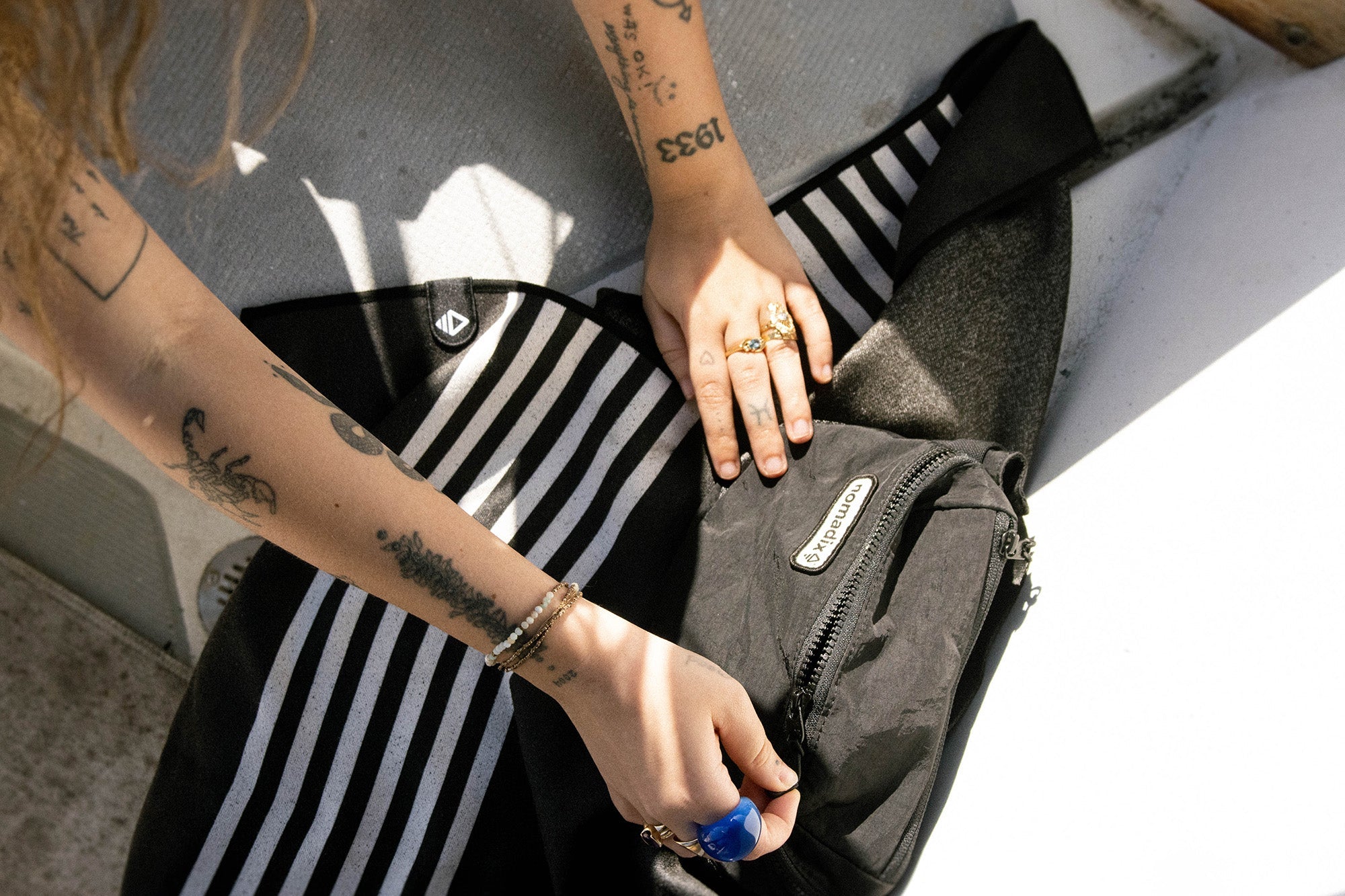 Tattooed person opening a black Nomadix backpack on black-and-white striped fabric in bright sunlight