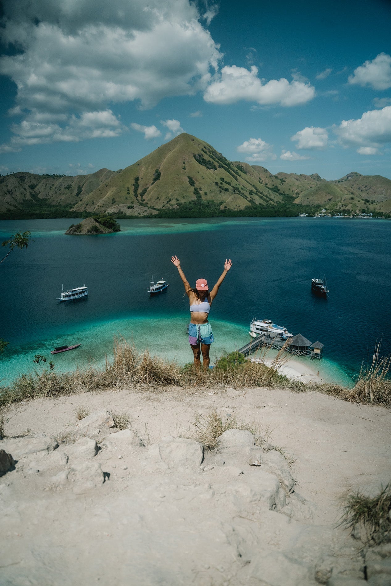 Woman with arms raised overlooking turquoise waters, boats, and rugged hills in Komodo National Park
