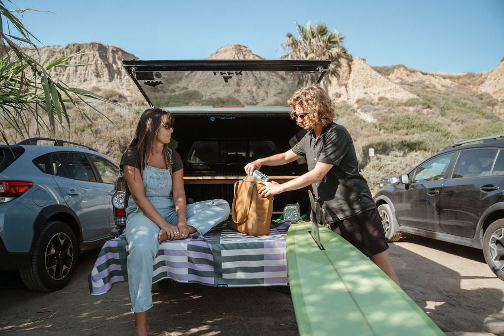 Two people prepare longboard surf gear beside a vehicle in a sunny rocky parking area