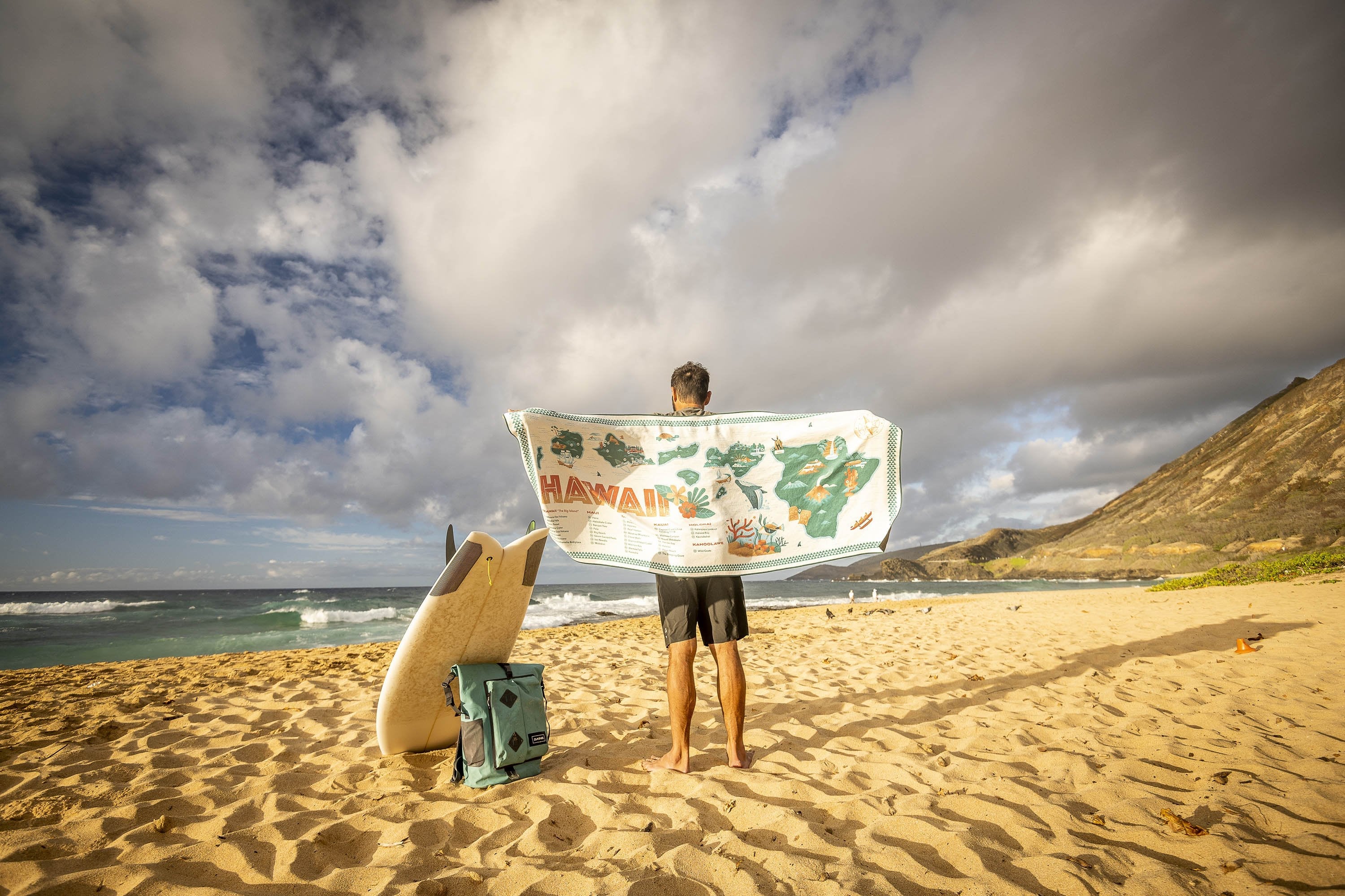 Person holding a Hawaii map towel on a beach beside a surfboard and backpack