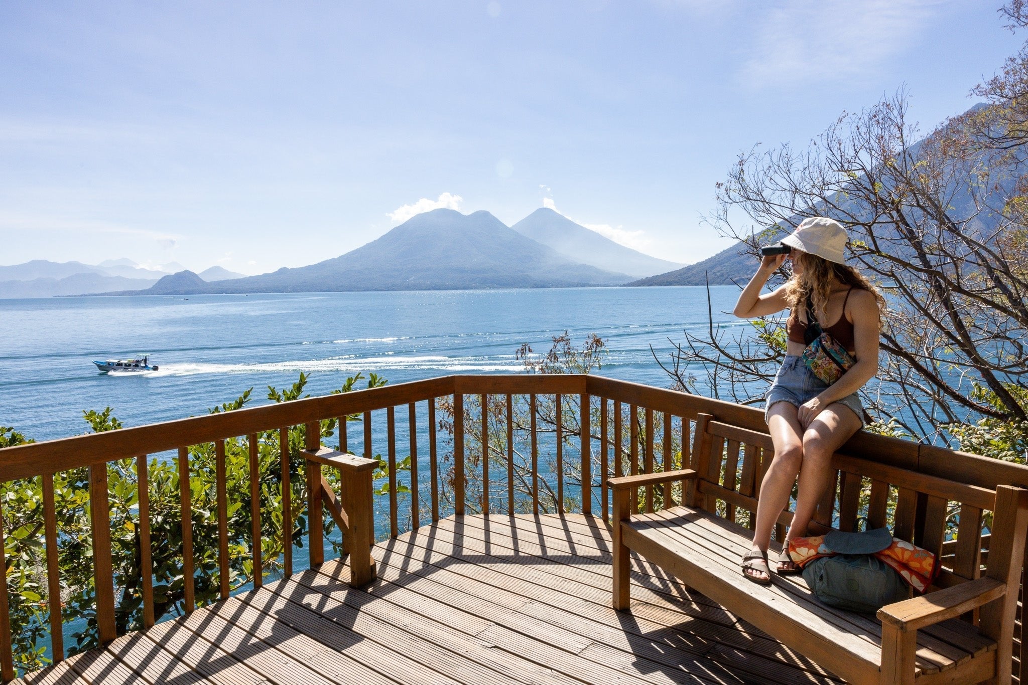 Woman looking through binoculars toward volcanic mountains across a lake from a wooden deck