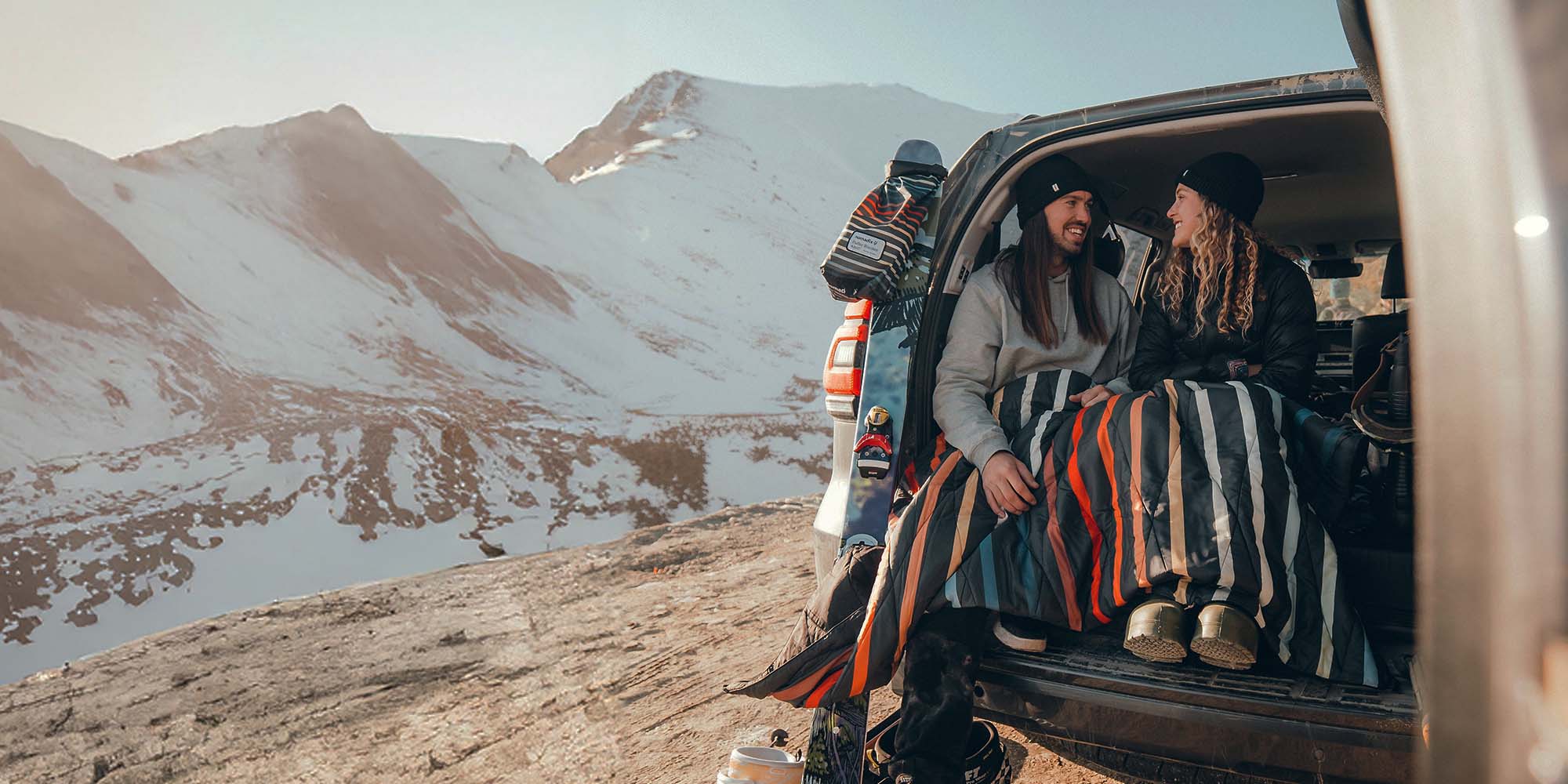 Two people sit in an open SUV with a striped blanket beside snow-covered mountains