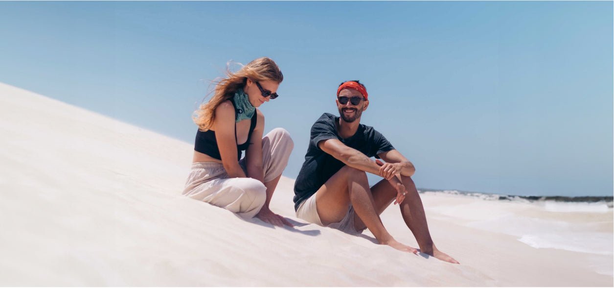 Two travelers sitting on a white Australian beach beside the ocean under a clear blue sky