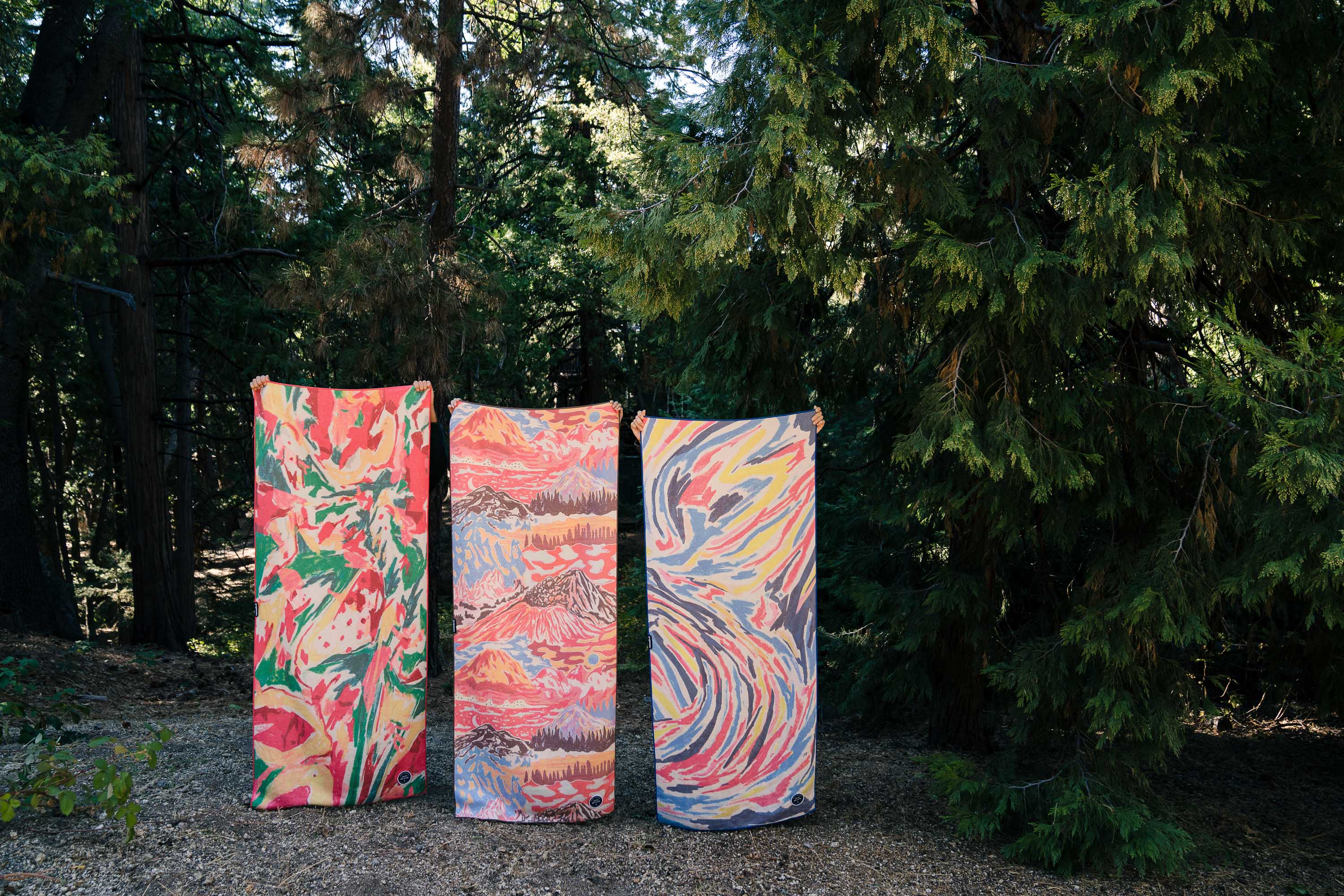 Front view of three Nomadix towels from the Jose Berrio collection showcasing colorful abstract patterns with a forest background