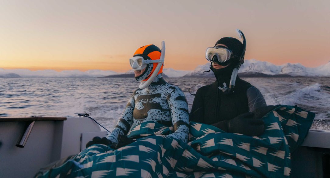 Two snorkelers wrapped in patterned blankets on a boat with snowy mountains across the Arctic sea