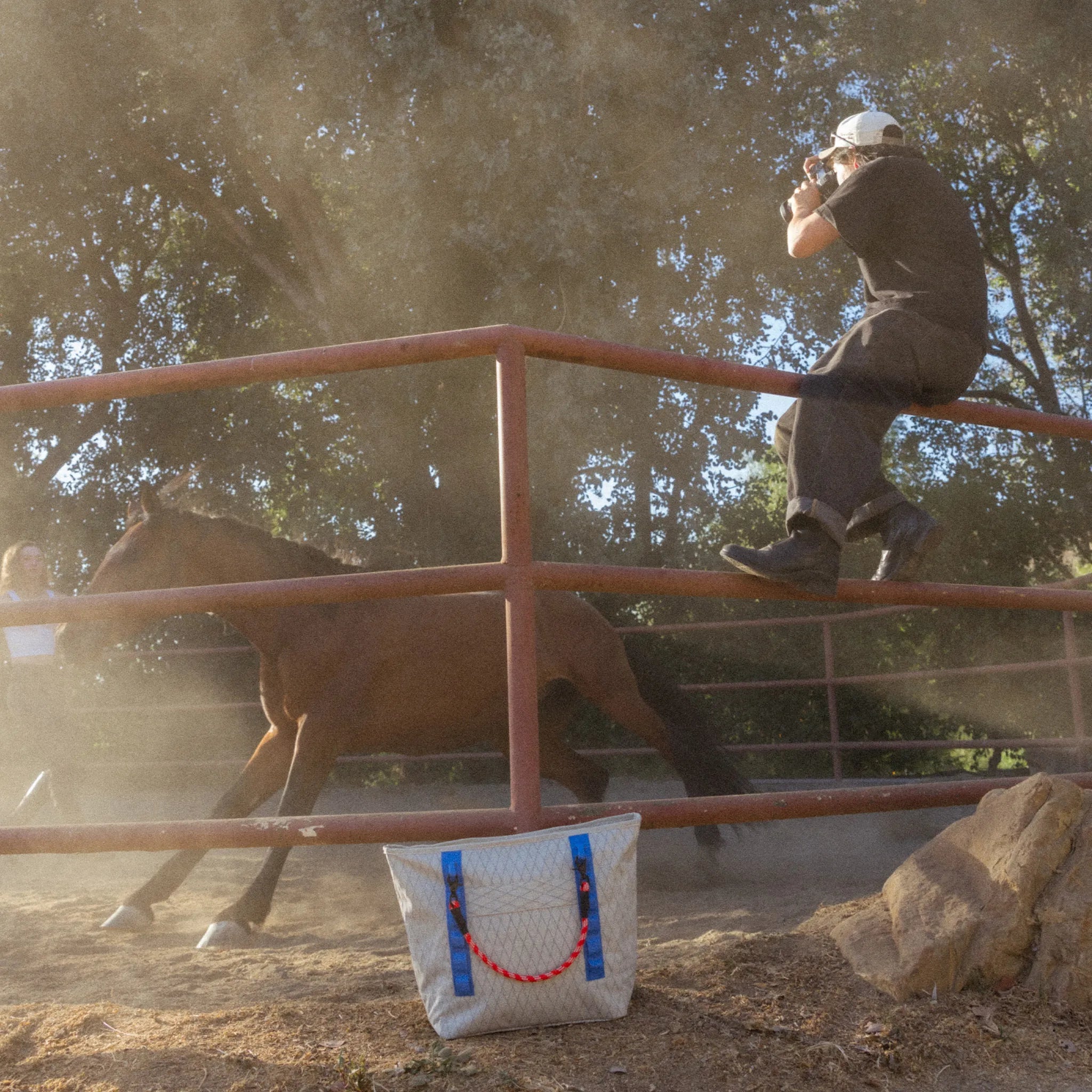 Person riding a horse over a rail in a dusty outdoor arena beside a white tote bag