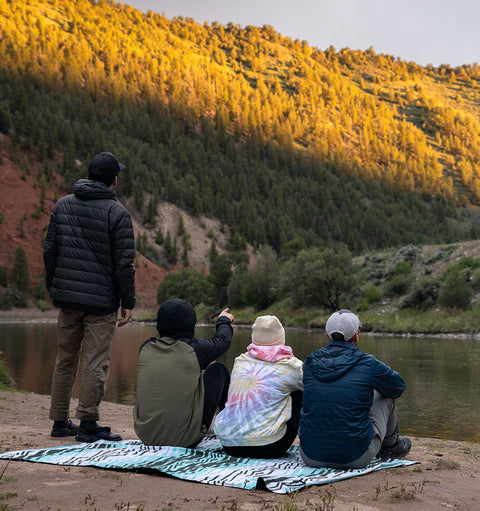Multiple people sit on a Nomadix Festival blanket while at the water's edge of a pond in the mountains.