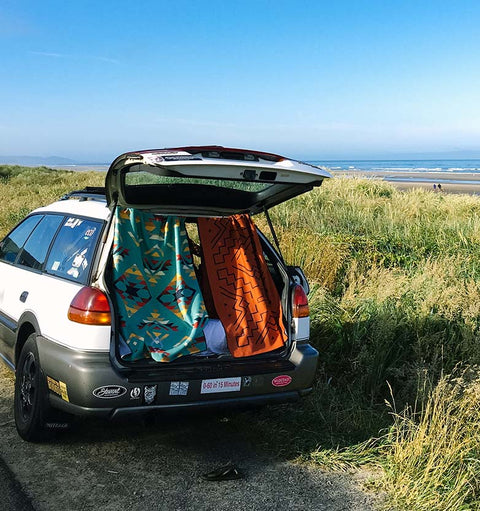 A white subaru car on the side of a road near a beach, with the back of the car open and two towels hanging down from the roof.