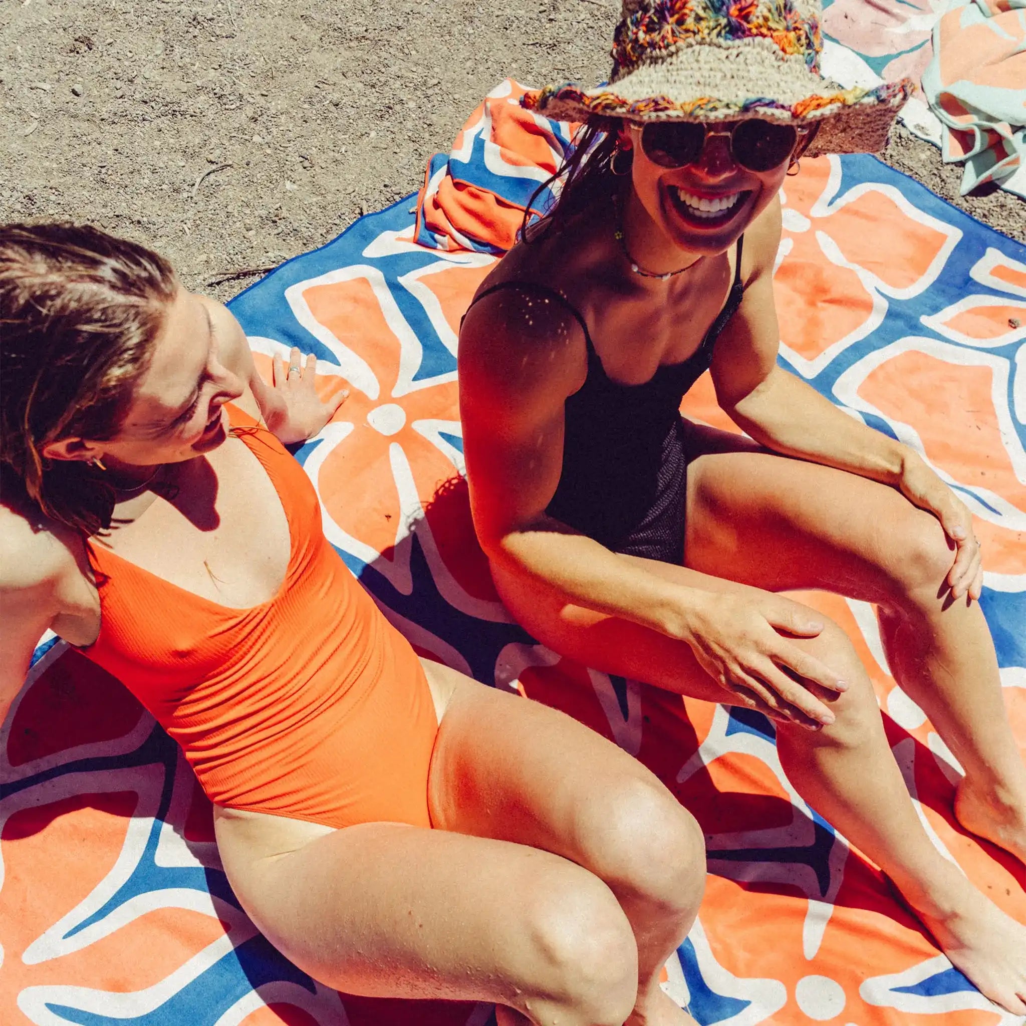 Two women relax on the Wild Flowers Stellar Blue Red blanket towel in a sunny sandy setting