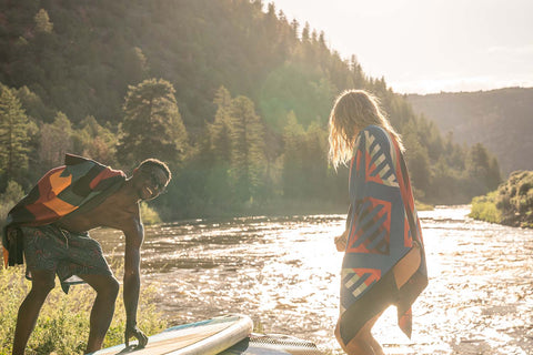 Two people get ready to paddleboard at the edge of a stream with Nomadix towels around them.