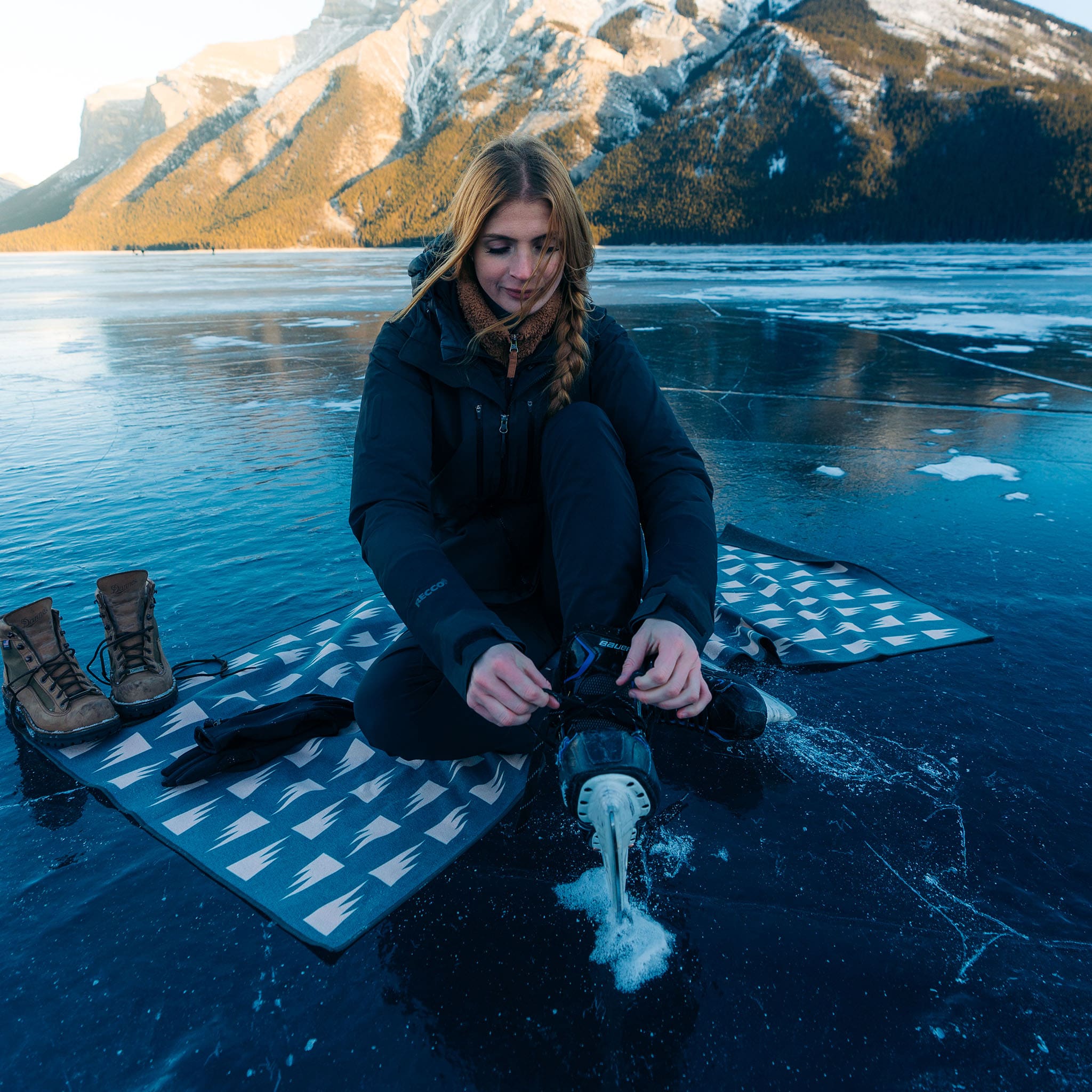 Woman adjusts an ice skate while sitting on the Summit Green Ultralight Towel on a frozen lake beneath mountains